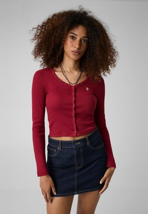 Donna con i capelli ricci che indossa un top rosso aderente a maniche lunghe con bottoni e una gonna di jeans blu scuro, in piedi davanti a uno sfondo monocromatico.