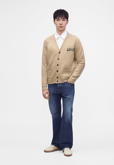 Jeune homme portant un cardigan beige boutonné avec un patch, une chemise blanche, un jean bleu et des baskets blanches, debout devant un fond uni.