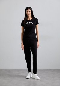 Svart bomull T-skjorte med hvitt EA7-logo, kombinert med svarte slim-fit bukser og hvite sneakers, stående mot en enkel grå bakgrunn.