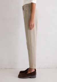 Beige cotton pants with a slim fit, featuring a mid-rise waist and classic back pockets. Worn with dark brown slip-on shoes.