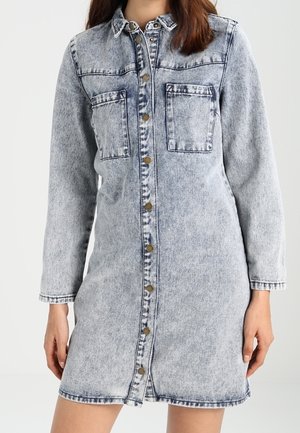 Kvinde iført en knappet, langærmet acid-vasket denimkjole med to brystlommer og krave, stående foran en ensfarvet baggrund.