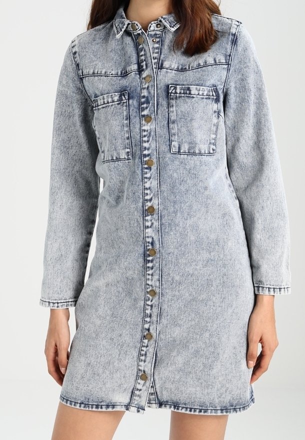 Femme portant une robe en jean acidulé, boutonnée, à manches longues, avec deux poches poitrine et un col, debout devant un fond uni.