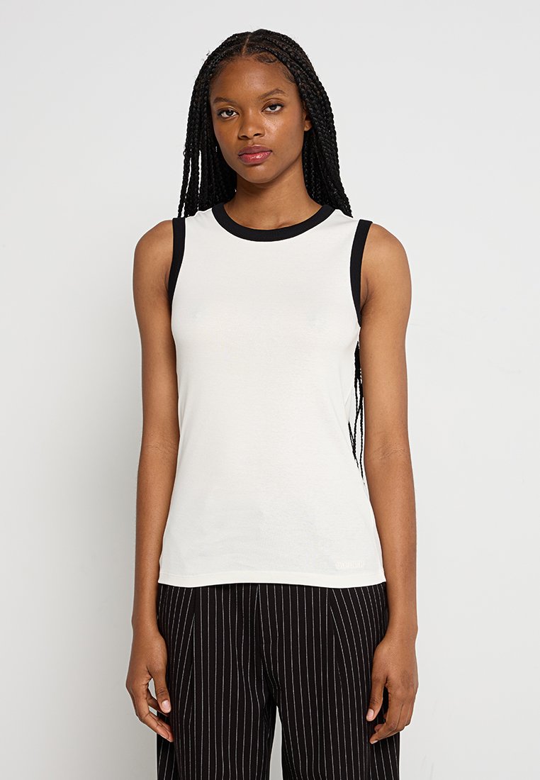 Biała, bezrękawnikowa bluzka z czarnym dekoltem i otworami na ramiona; miękka tkanina z dopasowanym krojem, noszona z czarnymi, paskowanymi spodniami.