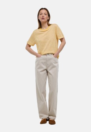 Femme debout, les mains dans les poches, portant une chemise jaune à rayures à manches courtes, un pantalon crème à fines rayures et des chaussures marron sans lacets.