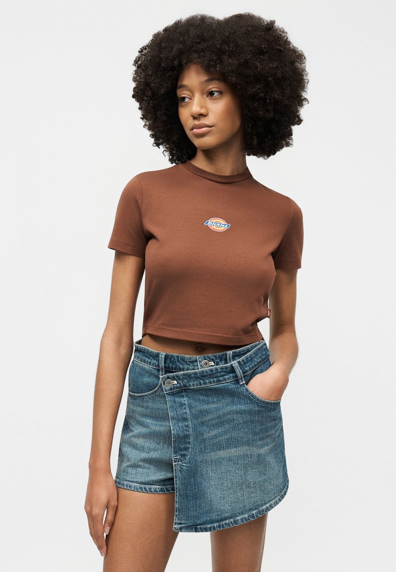 Donna con capelli naturali ricci che indossa una maglietta marrone corta con logo e una gonna di jeans asimmetrica con pannello frontale e tasche.