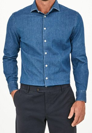 Mężczyzna w niebieskiej jeansowej koszuli z długim rękawem wsadzonej do ciemnoniebieskich spodni, z jedną ręką w kieszeni.