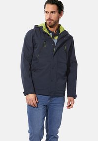 Navy blue jacket with a green hood, featuring two chest zip pockets and snap buttons. The material appears water-resistant with a smooth texture.