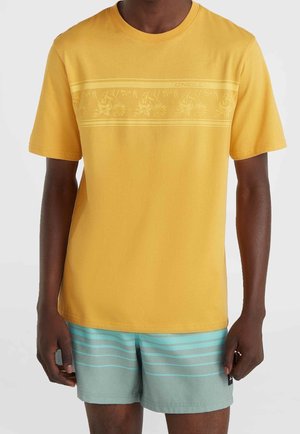 Homme portant un t-shirt jaune à manches courtes avec une discrète rayure florale et un short rayé bleu clair sur un fond uni.