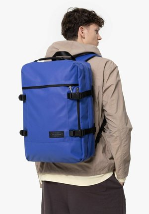 Zaino blu realizzato in tessuto resistente con spallacci neri, tasche con zip e forma rettangolare. Presenta una patch del logo frontale e clip laterali regolabili.