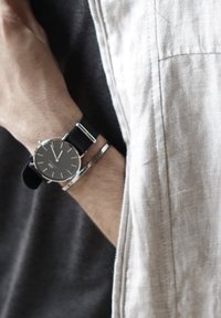 Orologio con cinturino nero e hardware argentato, quadrante rotondo, design minimalista, quadrante grigio e bracciale in argento al polso.