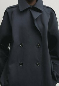 Granatowy płaszcz trench z podwójnym zapięciem, dużym kołnierzem i minimalistycznym designem, wykonany z gładkiego, strukturalnego materiału.