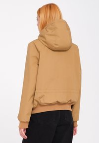 Khaki hooded jack met een zachte textuur, geribbelde mouwen en onderzoom. Heeft een relaxte pasvorm en minimale stiksels.