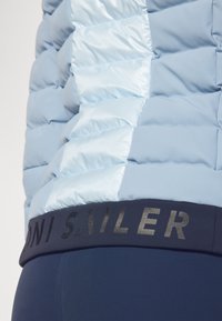 Hellblauer, gepolsterter Mantel mit glänzender Textur und gestepptem Muster, ausgestattet mit einem marineblauen Bund und schwarzem Text, der "MONI SAILER" liest.