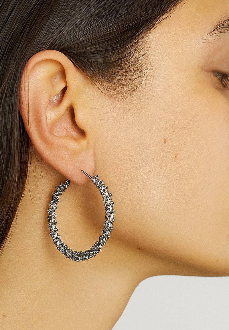 Silberne Creolen mit geflochtenem Design, featuring einer strukturierten Oberfläche und einem sicheren Verschluss, am Lappen getragen.