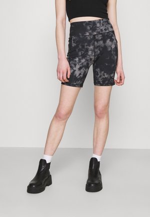 Kobieta ubrana w czarne i szare spodenki kolarskie tie-dye, czarny crop top, białe skarpety i czarne botki na platformie, stojąca na jednolitym tle.