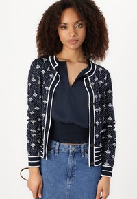 Navy cardigan with white floral patterns, featuring a button-up front and striped accents. Worn over a navy blouse and denim skirt.