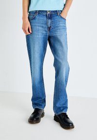 Person wearing light blue straight-leg jeans and black lace-up shoes, standing with one hand in pocket on white background.