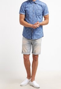 Camicia di jeans azzurro chiaro con maniche corte e tasca, abbinata a pantaloni corti chiari con effetto usurato e scarpe bianche senza lacci. Outfit estivo casual.
