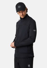Zwarte zip-up jas met textuurmouwen, voorzakken met rits en een hoge kraag. Heeft een logopatch op de linkermouw.