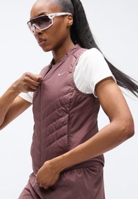 Καφέ γιλέκο τύπου puffer με καπιτονέ υφή και ψηλό γιακά, σε συνδυασμό με λευκό κοντομάνικο πουκάμισο. Ξεκάθαρα γυαλιά ηλίου ολοκληρώνουν την εμφάνιση.