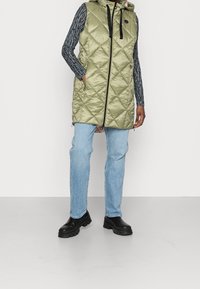 Kurtka pikowana w oliwkowym kolorze z kapturem i czarnymi akcentami, z zamkiem z przodu, kieszeniami bocznymi i długości do połowy uda. Połączona z jasnoniebieskimi dżinsami.