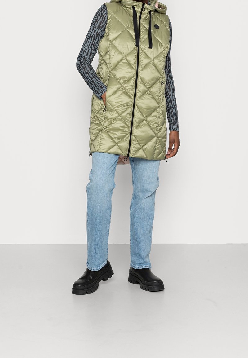 Kurtka pikowana w oliwkowym kolorze z kapturem i czarnymi akcentami, z zamkiem z przodu, kieszeniami bocznymi i długości do połowy uda. Połączona z jasnoniebieskimi dżinsami.