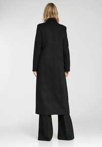 Długi czarny płaszcz o prostym kroju, teksturowana tkanina, kołnierz z wycięciem i długie rękawy, w połączeniu z szerokimi czarnymi spodniami.