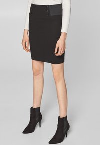 Černá tužková sukně vyrobená z elastického materiálu, s vysokým pasem a dekorativními knoflíky a hladkým povrchem. Doplněná černými kotníkovými botami.