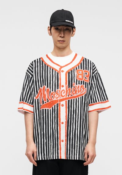 Πουκάμισο σε υπερμεγέθη γραμμή με ασπρόμαυρες ριγέ και πορτοκαλί λεπτομέρειες, με το κείμενο «Moschino». Κοντά μανίκια, κουμπιά μπροστά και χαλαρή εφαρμογή.
