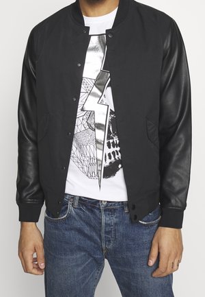Persona che indossa una giacca nera con maniche in pelle sopra una maglietta bianca con grafica che presenta un fulmine metallico e un design astratto, abbinata a jeans blu.