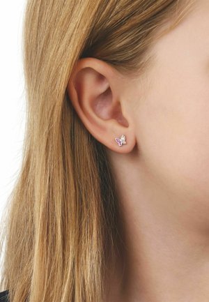 Gros plan sur l'oreille d'une femme aux longs cheveux blonds, portant une petite boucle d'oreille en forme de papillon avec des pierres roses et transparentes.