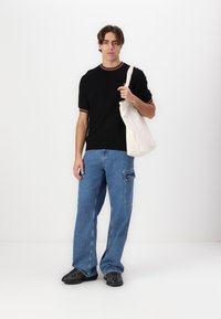 Black short-sleeve shirt with red and white ribbed collar, blue wide-leg jeans with side pocket, and black patterned slip-on shoes. Holds a beige tote bag.