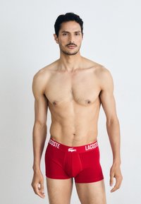 Red cotton boxer briefs with a white Lacoste logo on the waistband. The design is form-fitting with a smooth texture. Model is shirtless.