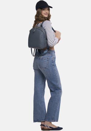 Grå læderrygsæk med dobbelte stropper, rundet form, sidelommer og gyldne accenter. Båret af en person i en stribet skjorte og jeans.