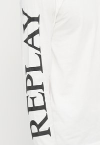 Fehér, hosszú ujjú ing, bal ujján függőlegesen a "REPLAY" felirattal, fekete színben. Puha anyag, standard szabás.