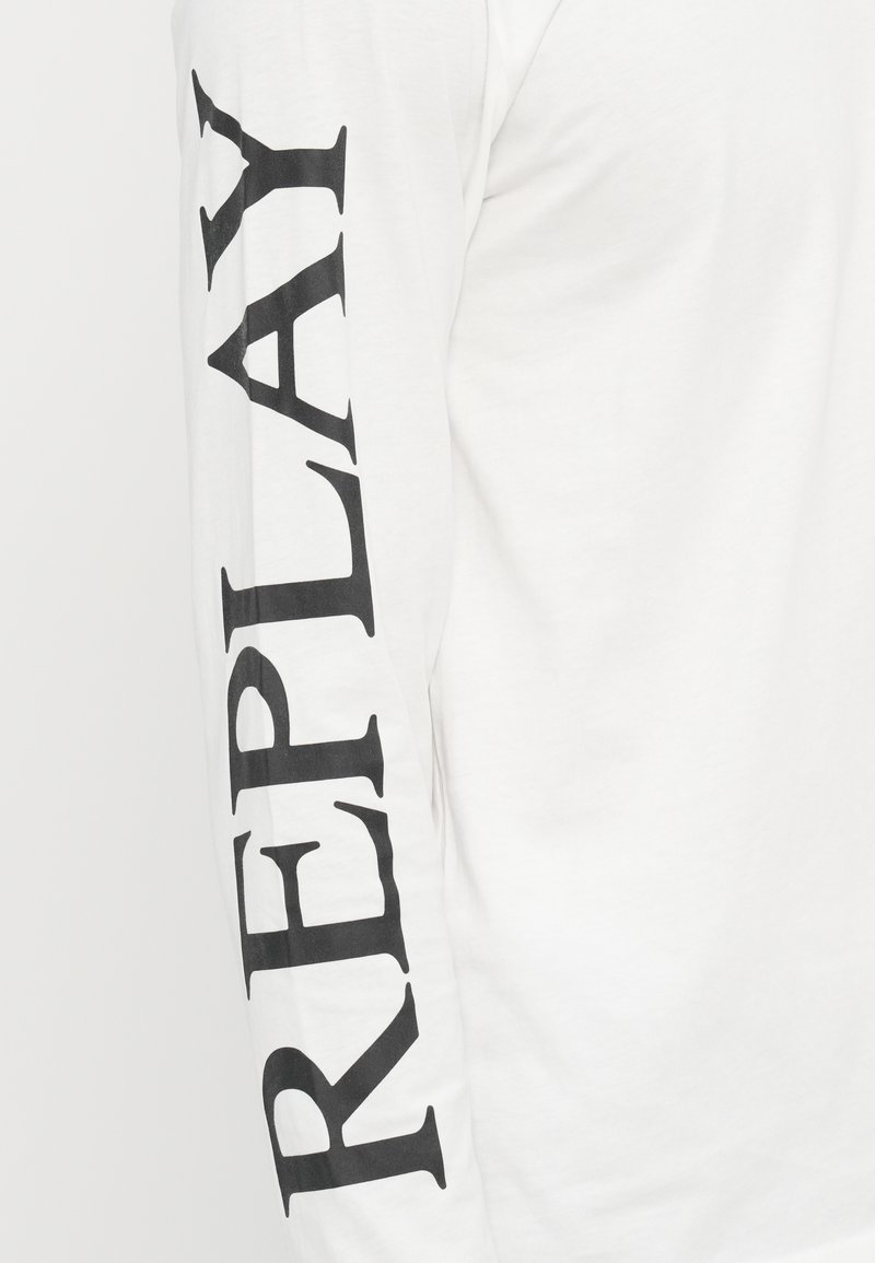 Camicia bianca a maniche lunghe con testo nero che legge "REPLAY" verticalmente lungo la manica sinistra. Tessuto morbido e vestibilità standard.