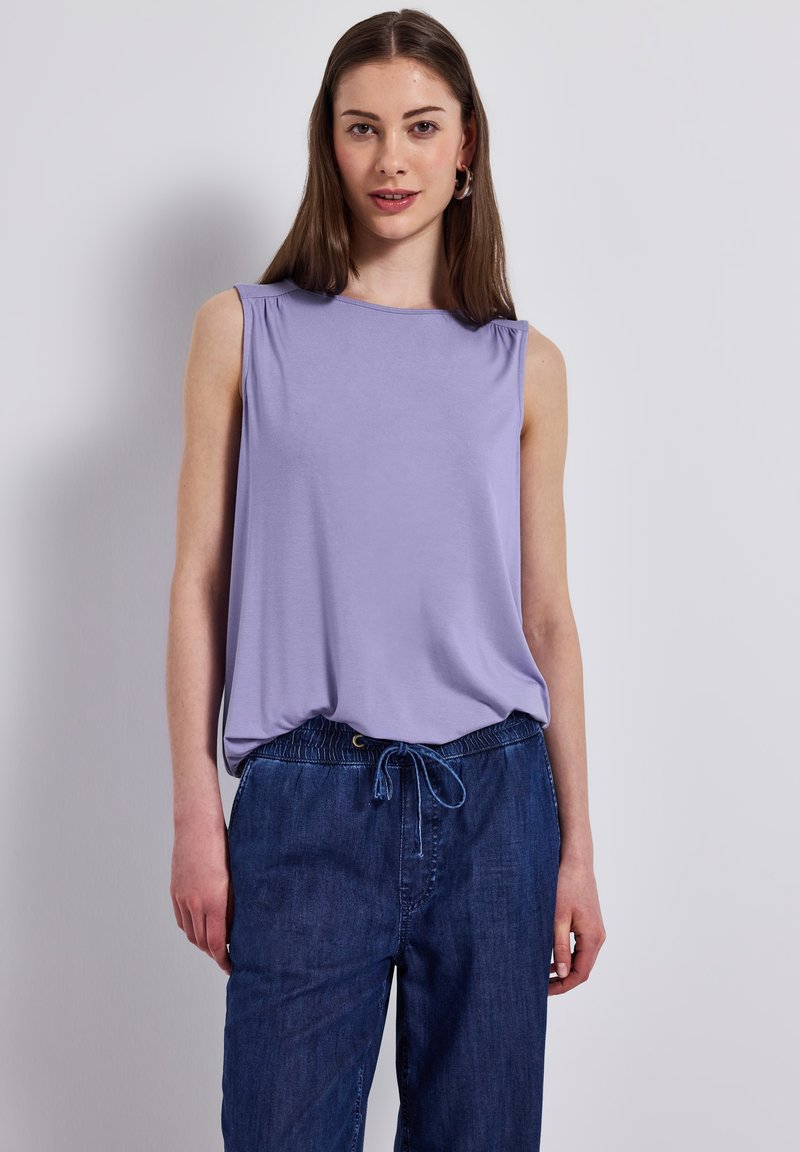 Top senza maniche di colore viola con vestibilità morbida, tessuto delicato e dettagli sottili sulle spalle, abbinato a pantaloni in denim elasticizzati di colore blu scuro.