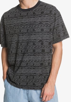 Man draagt een zwart t-shirt met grijze geometrische tribale patronen en lichtblauwe denim shorts, handen in zakken, tegen een witte achtergrond.