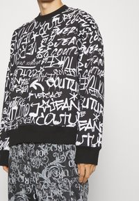Černá mikina s bílým textem ve stylu graffiti, kulatým výstřihem, dlouhými rukávy a žebrovanými man žetami. V kombinaci s odpovídajícími kalhotami se vzorem.