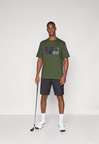 Mørkegrønn T-skjorte med svart grafisk trykk på forsiden, kombinert med svarte shorts, hvite sokker og sporty sko, mens han holder en golfklubbe.