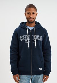 Sudadera con cremallera en color azul marino, con capucha forrada de forro polar, cordones blancos y bolsillos frontales. Impresión "CREEKS ORIGINAL Desde 1974" en el pecho.