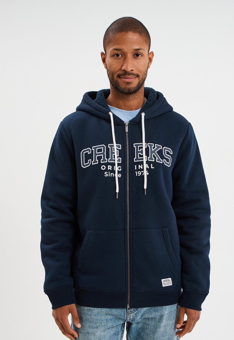 Sudadera con cremallera en color azul marino, con capucha forrada de forro polar, cordones blancos y bolsillos frontales. Impresión "CREEKS ORIGINAL Desde 1974" en el pecho.