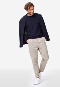 Pull marine et cardigan texturé sombre sur un pantalon beige clair. Baskets blanches. Le modèle se tient avec une main dans la poche, souriant.
