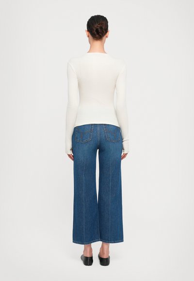 Long-sleeve white top paired with wide-leg blue denim jeans. Features two back pockets and a straight hem, minimal stitching details.