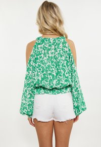 Blusa verde con motivo floreale, con maniche lunghe e svolazzanti e un colletto arricciato, abbinata a pantaloni corti in denim bianco strappato.
