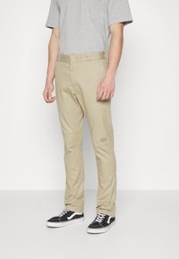 Man wearing beige straight-leg pants, gray short-sleeve t-shirt, and black and white sneakers, standing against a white background.