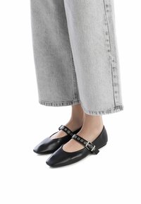 Bailarinas de cuero negro con punta afilada, que cuentan con una correa de tobillo con tachuelas y un diseño elegante, combinadas con jeans cortos grises.