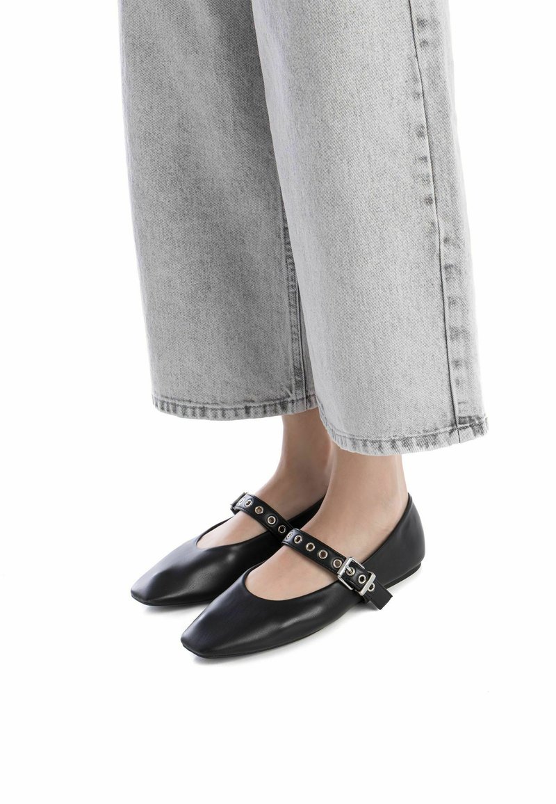 Bailarinas de cuero negro con punta afilada, que cuentan con una correa de tobillo con tachuelas y un diseño elegante, combinadas con jeans cortos grises.