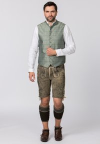 Traditionelle Tracht mit einer grünen gemusterten Weste, weißem Hemd, braunen mit Stichen verzierten Lederhosen, knielangen dunklen Socken und braunen Schuhen.