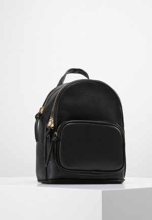Kleiner schwarzer Rucksack mit goldenen Reißverschlüssen, Fronttasche und oberem Tragegriff, auf einer weißen Oberfläche vor einfachem Hintergrund dargestellt.